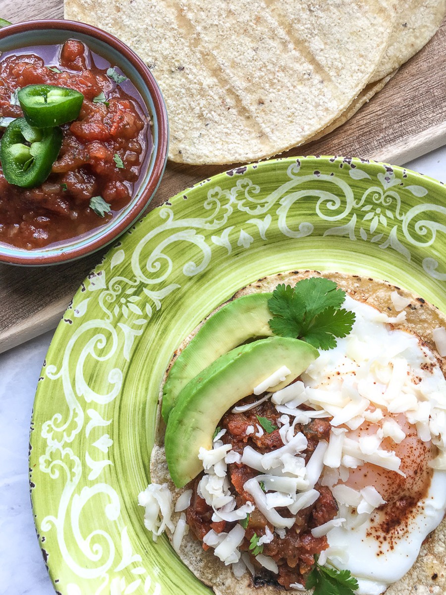 huevos rancheros on a plate topped with salsa, refried beans, shredded cheese and avocado slices.