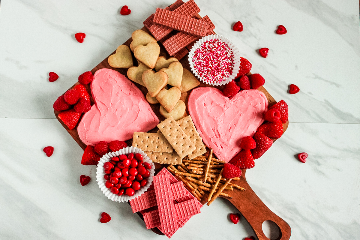 A valentine’s day dessert charcuterie board filled with heart-shaped cookies, pink frosting to dip them in, fresh fruit and candy.