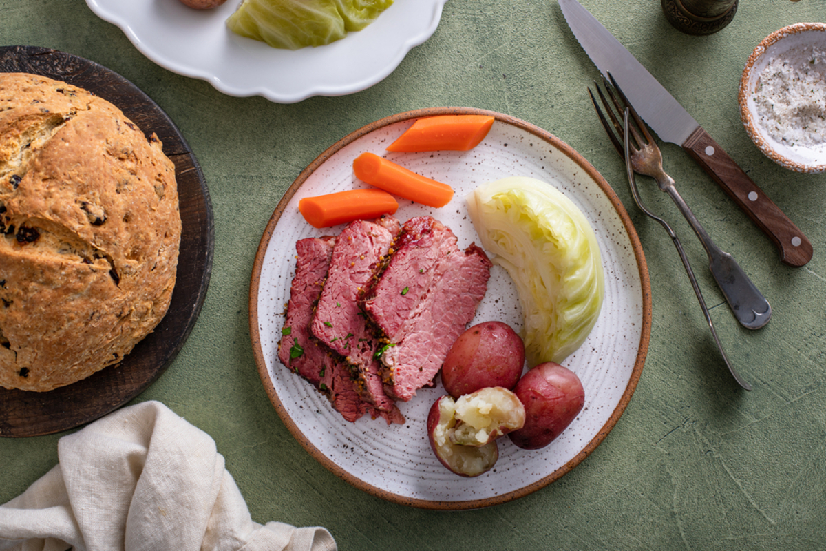 slices of slow cooker corned beef, carrots, cabbage and red potatoes on a plate with a loaf of irish soda bread on a cutting board